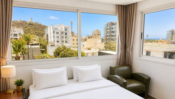 A hotel room with a bed, a chair, a lamp, and a view of buildings and a hill.