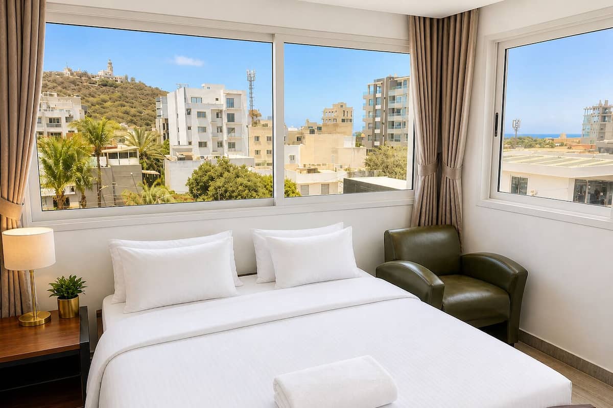 A hotel room with a bed, a chair, a lamp, and a view of buildings and a hill.