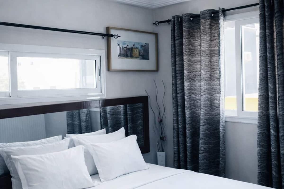 A bedroom with a bed, white bedding, a framed picture on the wall, and a window with curtains.