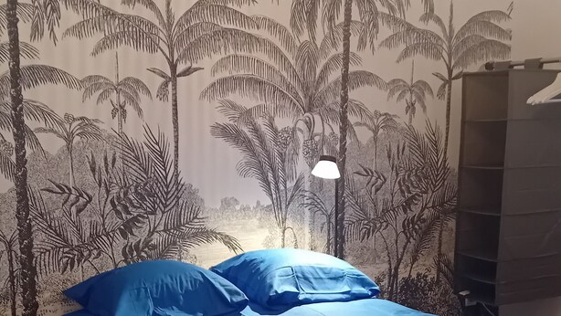 A bed with blue bedding, a wall with a palm tree mural, and a hanging light fixture.