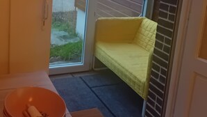 A room with a yellow cushioned bench, a wooden table with a bowl and a bottle, and a view of a house through a glass door.