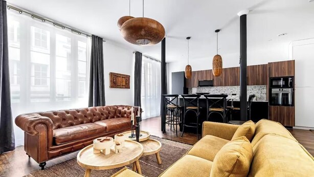 Un salon moderne avec un canapé en cuir marron, un fauteuil jaune et une table basse en bois. On y trouve également un coin cuisine avec un ensemble de salle à manger et des luminaires suspendus.