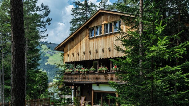 A wooden chalet with a balcony, surrounded by lush greenery and a mountainous backdrop. - B&B Villa La Bercia (Marebbe)
