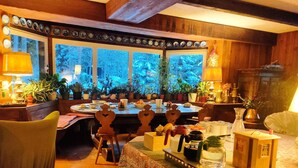 A cozy dining area with wooden furniture, a patterned tablecloth, and a view of the forest through large windows. - B&B Villa La Bercia (Marebbe)