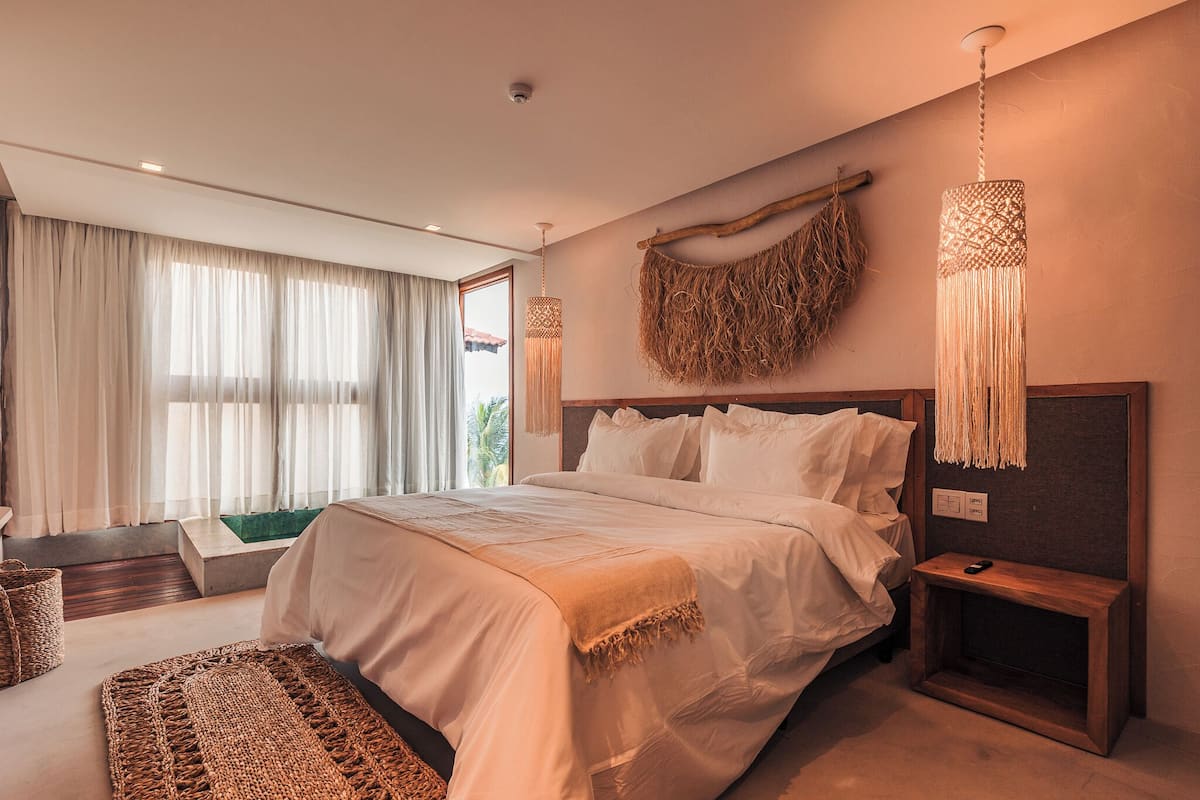 Une chambre à coucher moderne avec un grand lit, une table de chevet en bois, une décoration murale en fibres tressées et une vue sur l’extérieur.