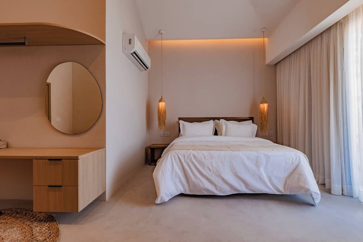 Une chambre moderne avec un grand lit, un miroir rond, un bureau en bois et deux lampes suspendues.