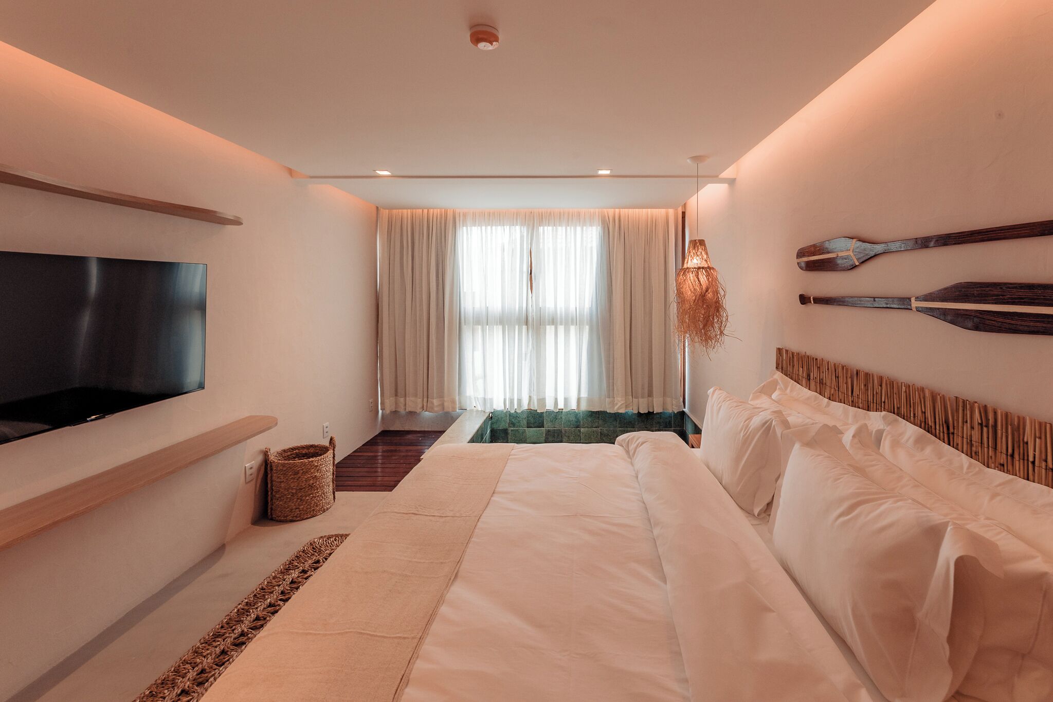 A modern hotel room with a large bed, a flat-screen TV mounted on the wall, a small seating area with a woven basket, and a view of the outdoors through sheer curtains.
