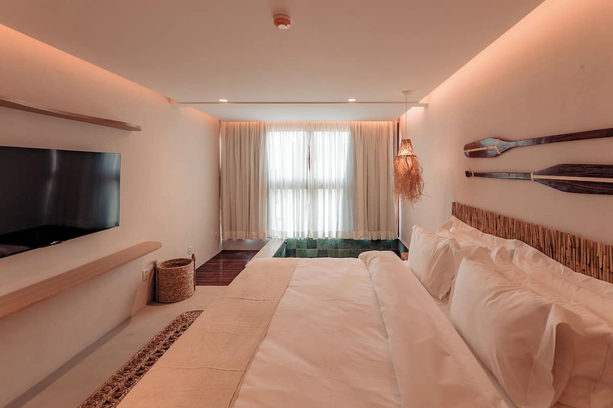 Une chambre d’hôtel moderne dotée d’un grand lit, d’un téléviseur à écran plat fixé au mur, d’un petit coin salon avec un panier en osier, et offrant une vue sur l’extérieur à travers des rideaux légers.