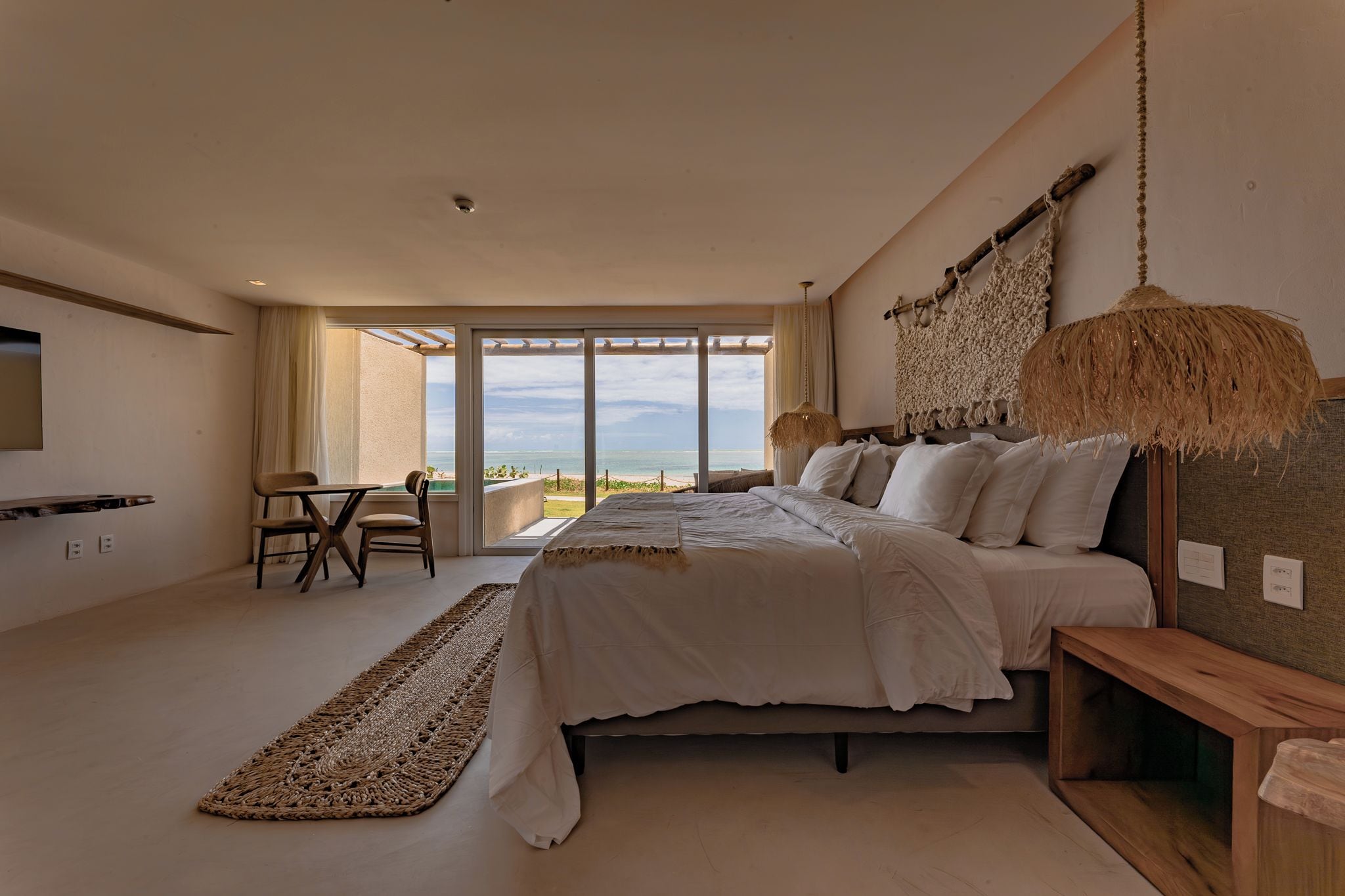 A bedroom with a large bed, a view of the sea, a dining area, and a TV mounted on the wall.