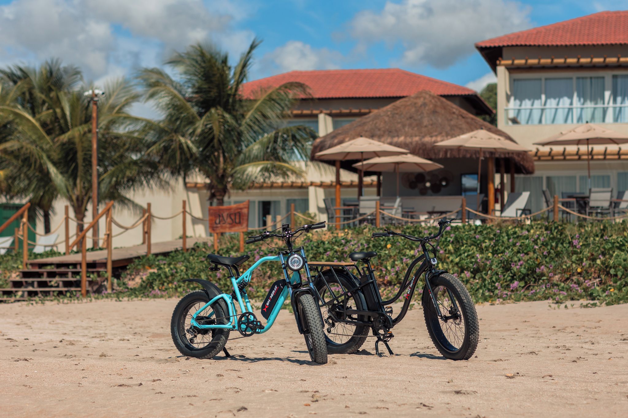 Ein türkisfarbenes Mountainbike mit Gepäckträger steht auf sandigem Untergrund, im Hintergrund ein Resort und Palmen.