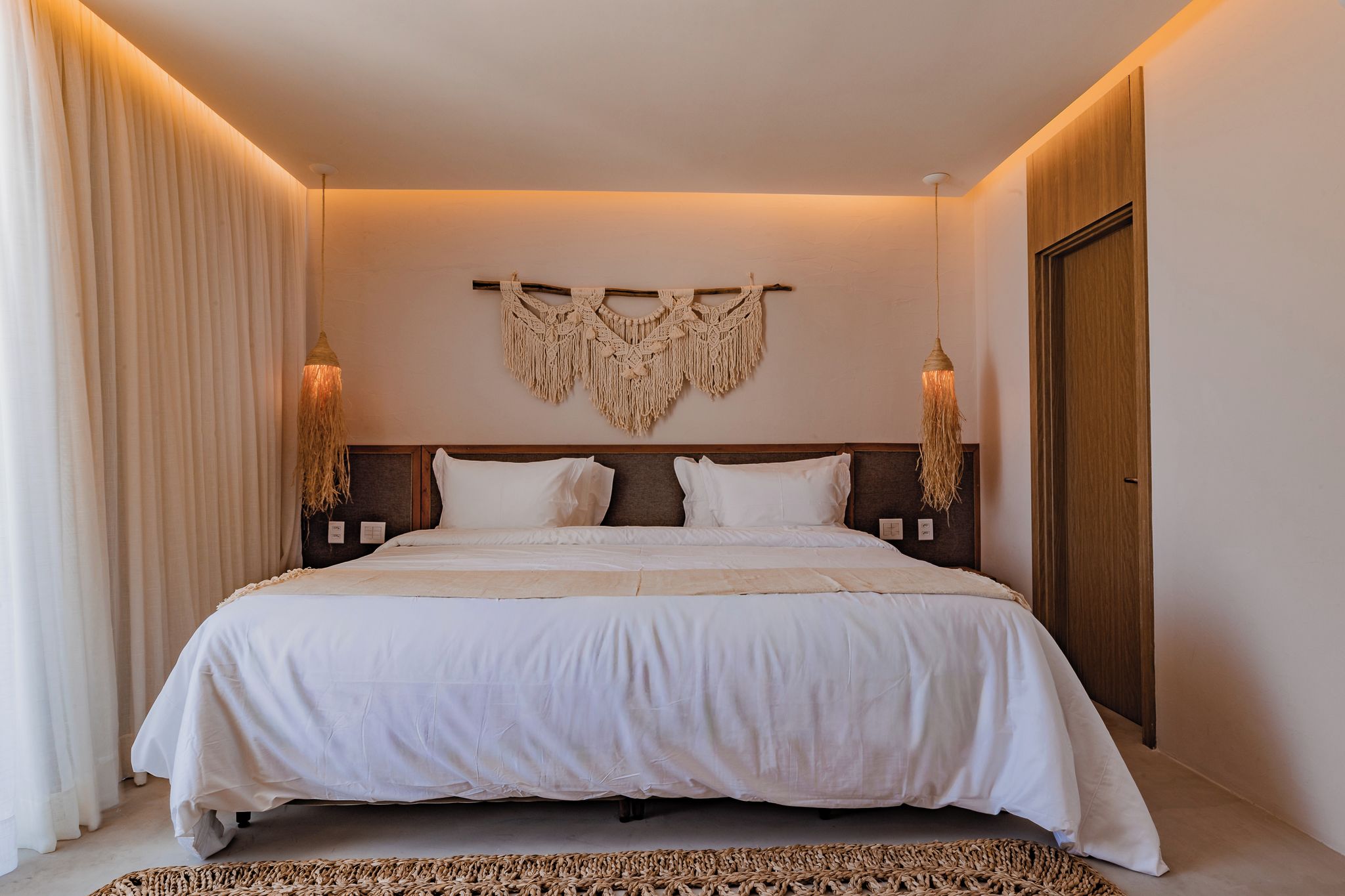 Une chambre d’hôtel avec un grand lit, deux tables de chevet, une décoration murale en macramé et une porte en bois.