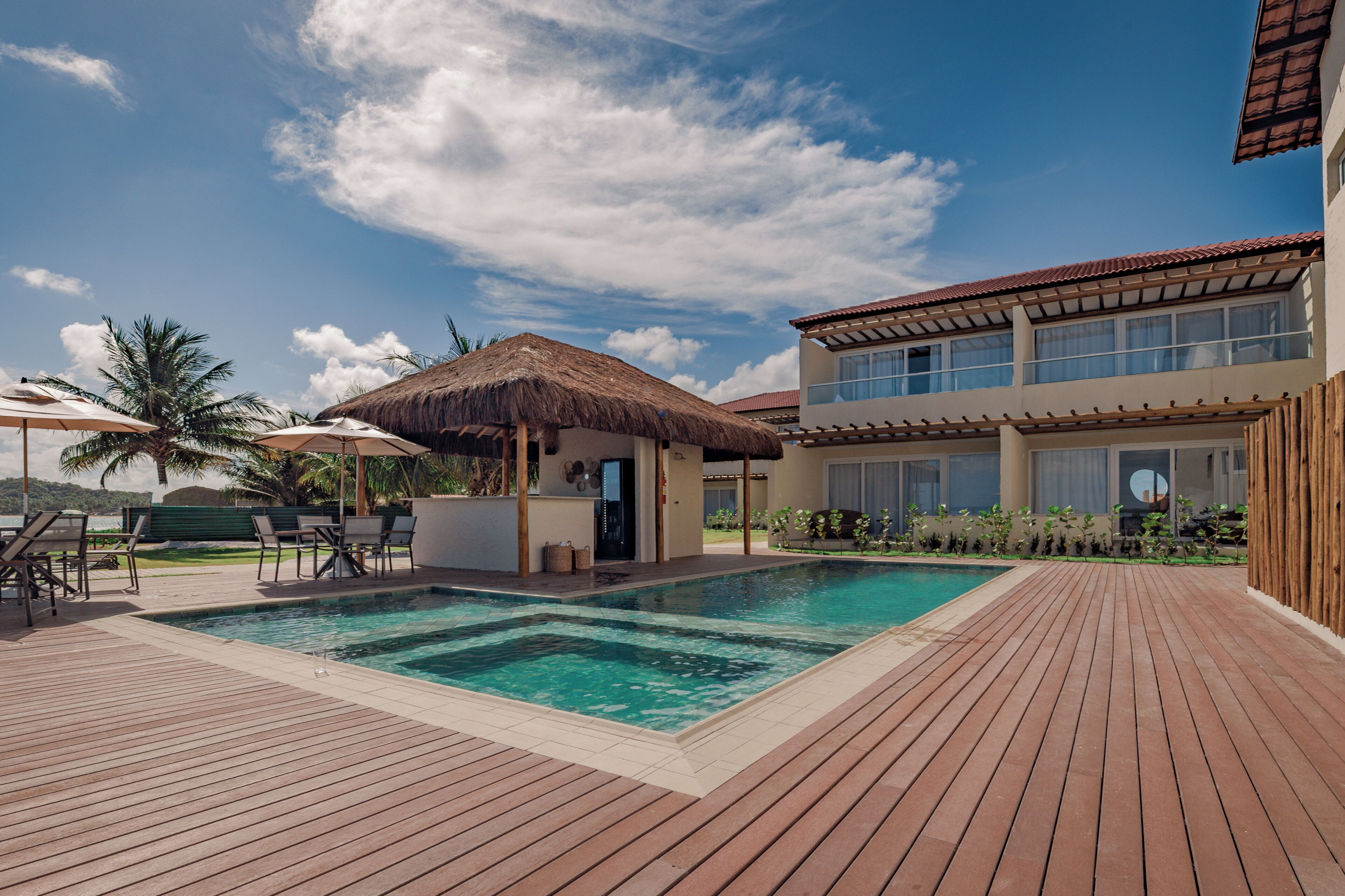 Una piscina con muelle de madera, una estructura cubierta de paja y un edificio moderno con amplios ventanales.