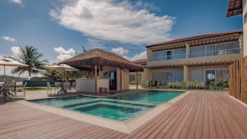 Una piscina con muelle de madera, una estructura cubierta de paja y un edificio moderno con amplios ventanales.