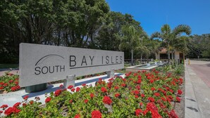 Skilt med teksten 'South Bay Isles', et anlagt område med røde blomster og en mindre bygning i baggrunden.