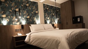 Une chambre d’hôtel avec un grand lit, une tête de lit en bois et un mur recouvert d’un papier peint à motifs floraux. On y trouve une table de chevet avec un téléphone et une commode sur laquelle est posée une télévision.