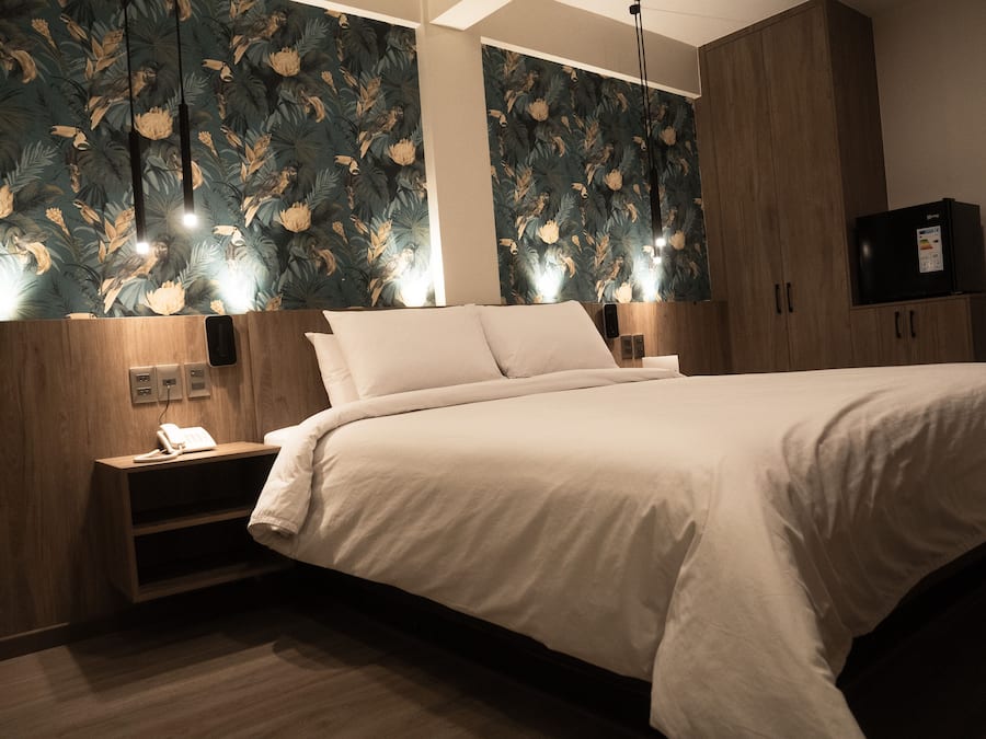 A hotel room with a large bed, wooden headboard, and a wall with floral wallpaper. There is a nightstand with a phone and a cabinet with a TV.