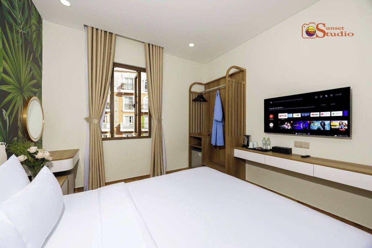 Ein Hotelzimmer mit einem Bett, einem an der Wand befestigten Fernseher, einem Fenster mit Vorhängen, einem Holzschrank und einem kleinen Tisch mit einer Blumenvase.
