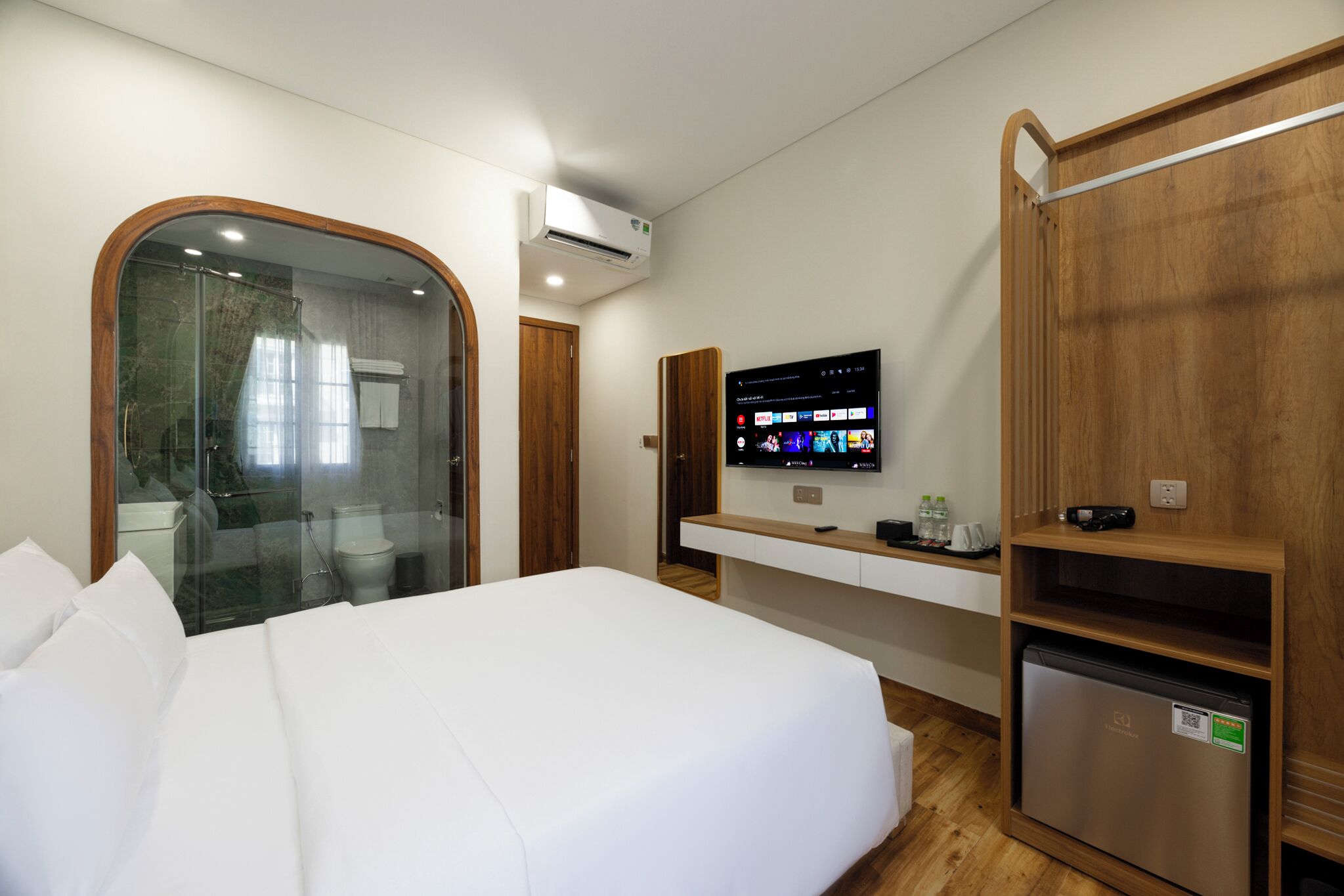 A hotel room with a bed, a TV mounted on the wall, a small fridge, and a wooden wardrobe.