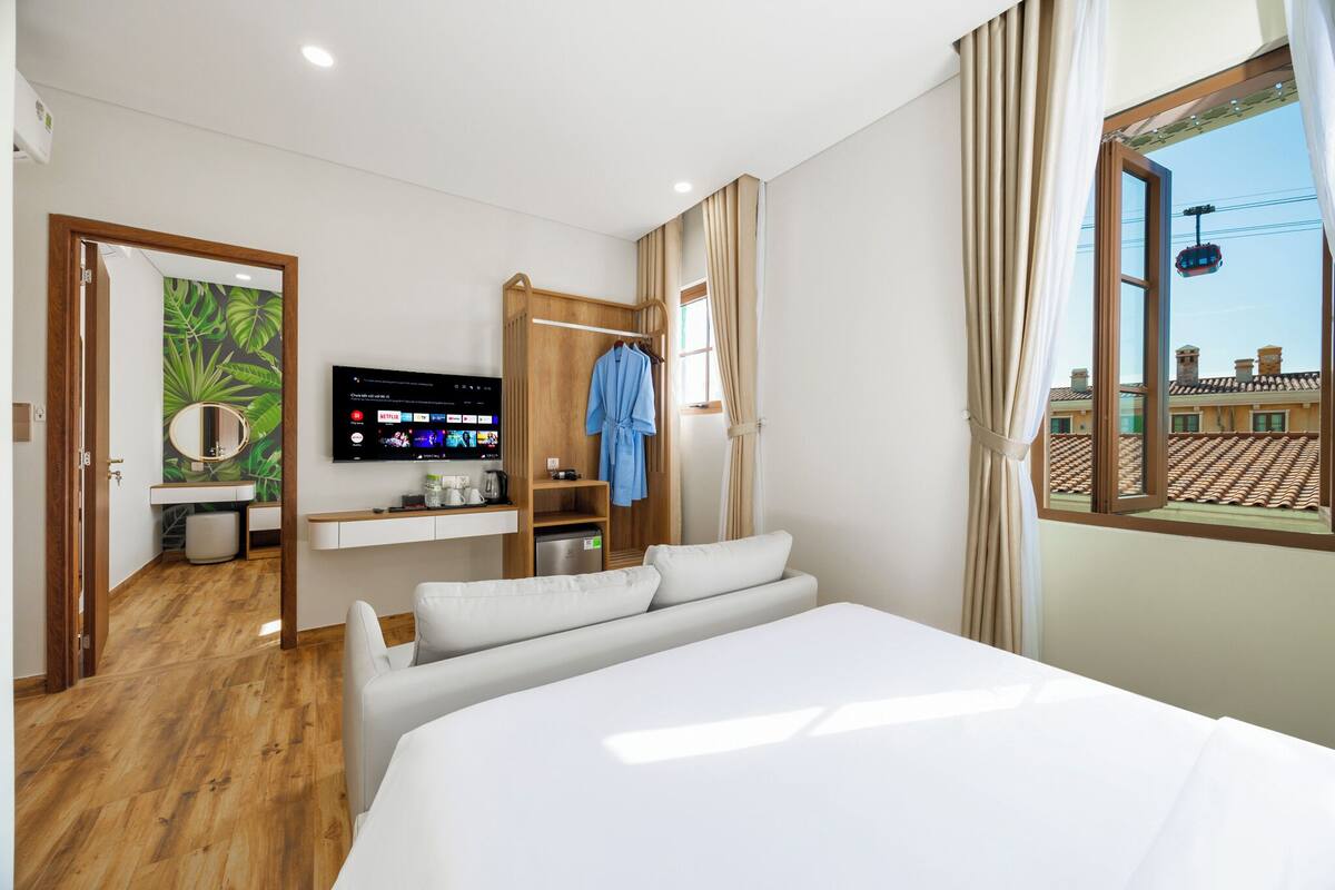 Ein Hotelzimmer mit Bett, Fernseher, kleinem Schreibtisch und Fenster mit Blick auf Gebäude.