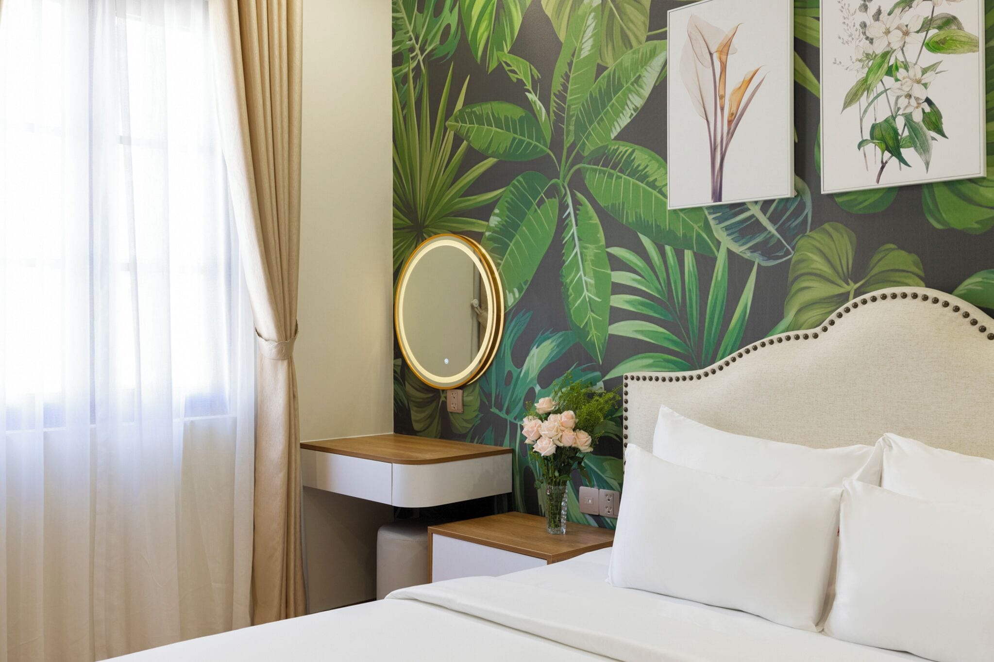 A hotel room with a bed, a bedside table with a mirror, a small table with a vase of flowers, and a wall with tropical wallpaper.