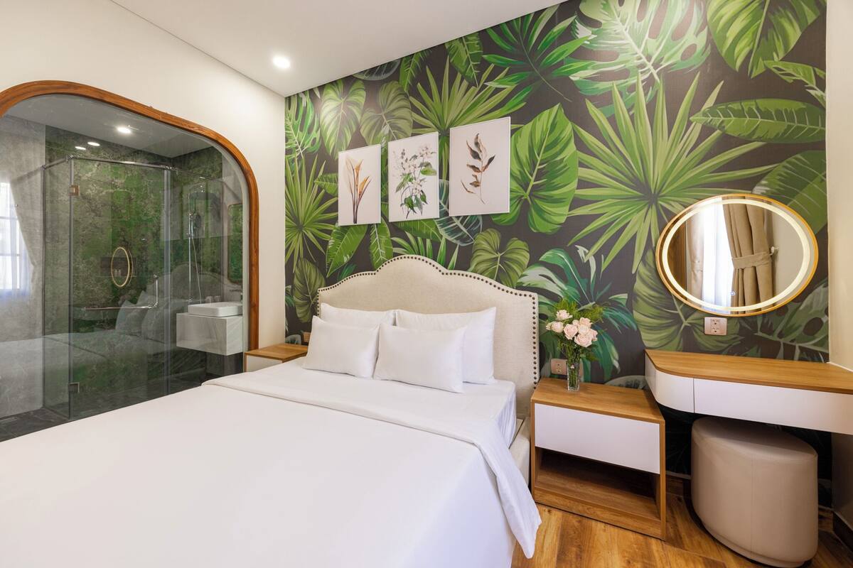 Ein modernes Hotelzimmer mit einer gläsernen Duschkabine, einem großen Bett mit weißer Bettwäsche, einem Nachttisch aus Holz mit Spiegel und einer Wand mit tropischem Blattmuster.