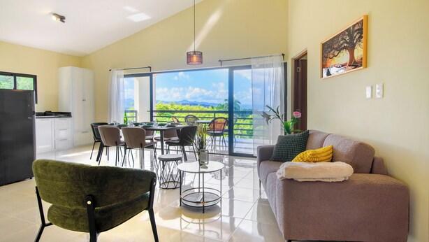 A modern living room with a sofa, chairs, a dining table, and a large window offering a view of greenery. - Casa Vista Mar 3 (Rio San Juan)