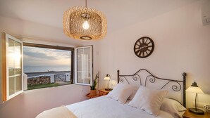 A bedroom with a bed, a window with an open view, a wall clock, and a hanging light fixture.