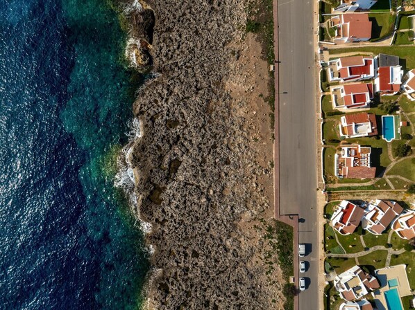 An aerial view of a coastal residential area with houses, a road, and a rocky shoreline.