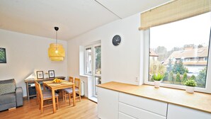 Una cocina moderna con suelo de madera, armarios blancos, un área de comedor con mesa y sillas, una lámpara colgante y una ventana con vistas al barrio.