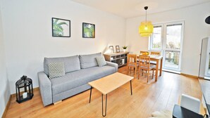Una sala de estar moderna con un sofá gris, una mesa de centro de madera, y un comedor con una mesa y sillas de madera. Hay dos cuadros en la pared, una planta pequeña en una mesita auxiliar, y una lámpara colgante amarilla.