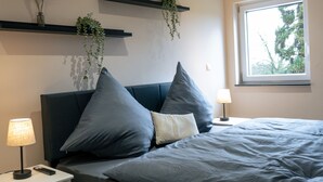 Une chambre à coucher comprenant un lit, des tables de chevet, une fenêtre et des étagères murales ornées de plantes.