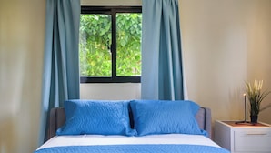 A bedroom with a bed, curtains, a window with a view of greenery, a nightstand with a plant, and a drawer. - Casa Vista Mar 4 (Rio San Juan)