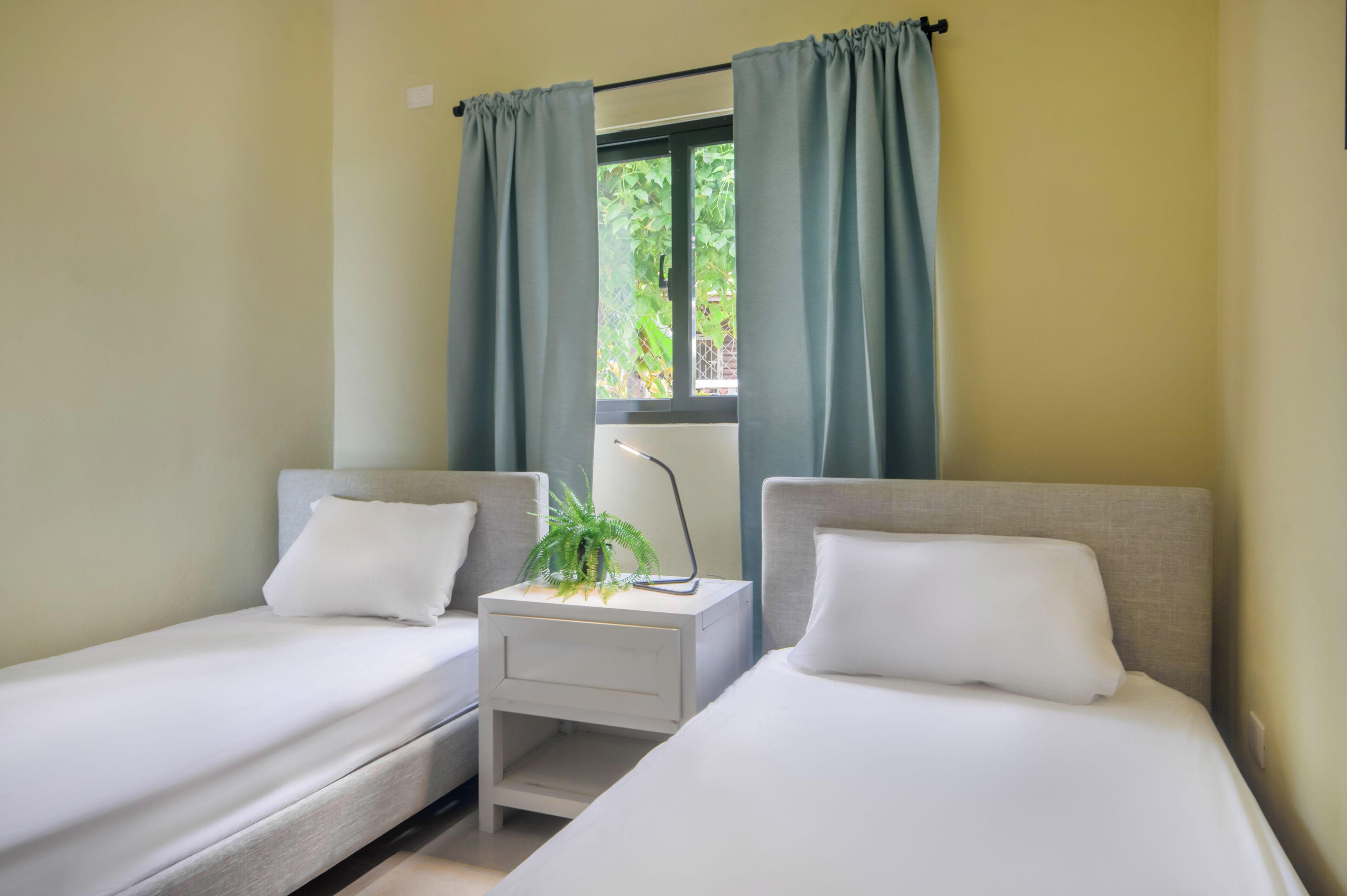 Habitación doble con dos camas individuales, una mesita de noche pequeña con lámpara y una ventana con cortinas.