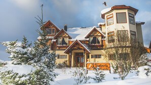 A chalet-style hotel with a snow-covered roof and multiple windows, surrounded by snow and trees.