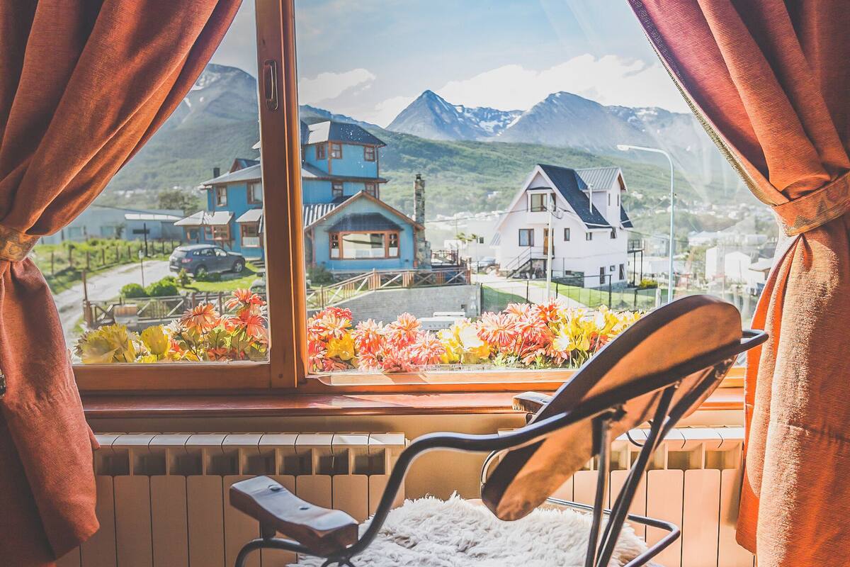 Une pièce avec vue sur un paysage montagneux, un fauteuil doté d’un coussin moelleux et une fenêtre avec des rideaux.