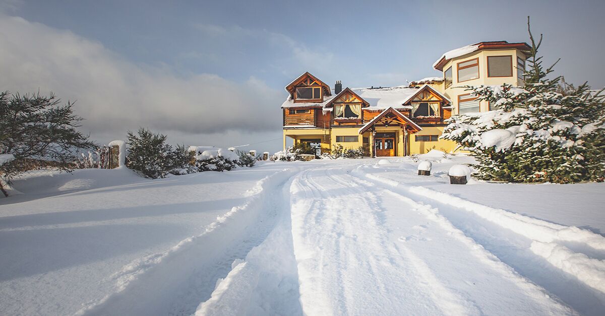 A large, multi-story wooden house with a snow-covered driveway and surrounding trees.