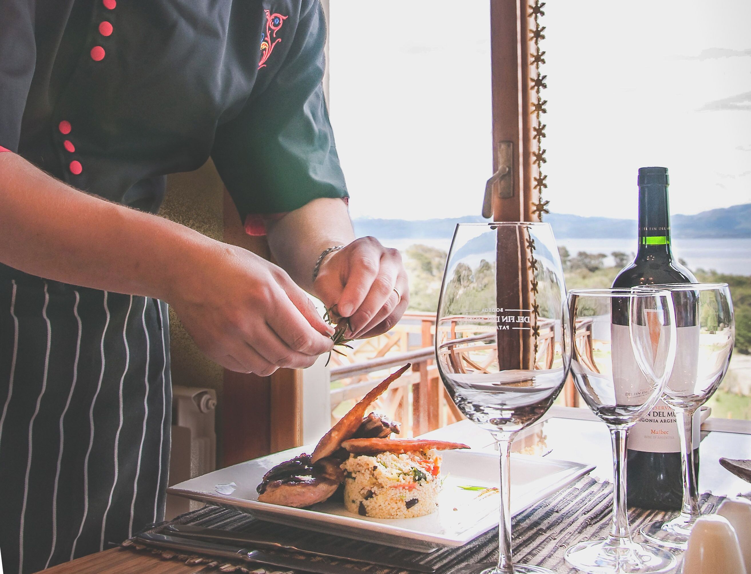 Un chef preparando comida sobre una mesa con una botella de vino y copas.