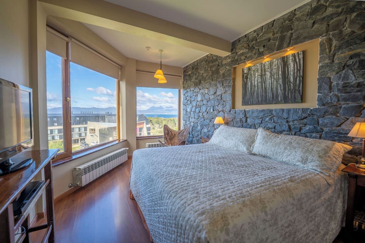 Une chambre avec un grand lit, un mur en pierre et une vue sur des immeubles et des montagnes.