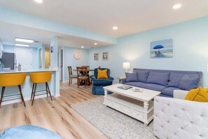 A living room with a sofa, coffee table, and chairs, a dining area with a table and chairs, and a kitchen area with a counter and stools.