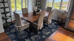 Un comedor con mesa de madera y sillas de mimbre, ventanas amplias con vista al océano, y una alfombra con estampado de estrella de mar.