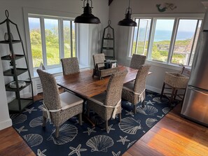 A dining area with a wooden table and wicker chairs, large windows with a view of the ocean, and a starfish-patterned rug. - Charming Handicapped Accessible 3-bedroom house in Welcoming Wellfleet (Wellfleet)