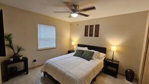 A bedroom with a bed, two bedside tables, a ceiling fan, a window with blinds, and three wall-mounted decor pieces.