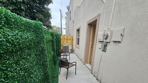 A narrow alley with a green hedge on one side and a building with a door and windows on the other. There are two chairs and a table in the alley.