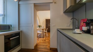 Une cuisine moderne équipée d’un four à micro-ondes, d’un four et d’une machine à café. On aperçoit un coin repas avec une table et des chaises à travers une porte ouverte.