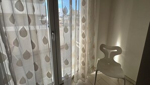 Une pièce dotée d’une porte-fenêtre coulissante, d’un rideau transparent à motifs de feuilles, d’une chaise blanche et d’une vue sur les bâtiments environnants.
