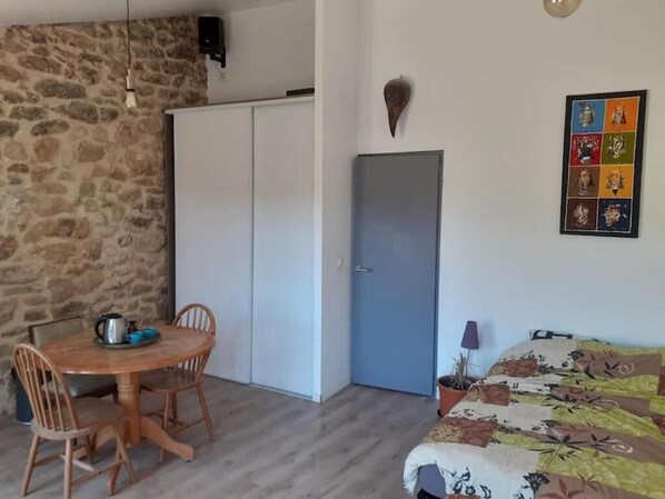 A room with a stone wall, a bed with a floral bedspread, a dining table with chairs, a blue door, and a framed pop art piece on the wall. - Old restored village house with terrace (Jouques)