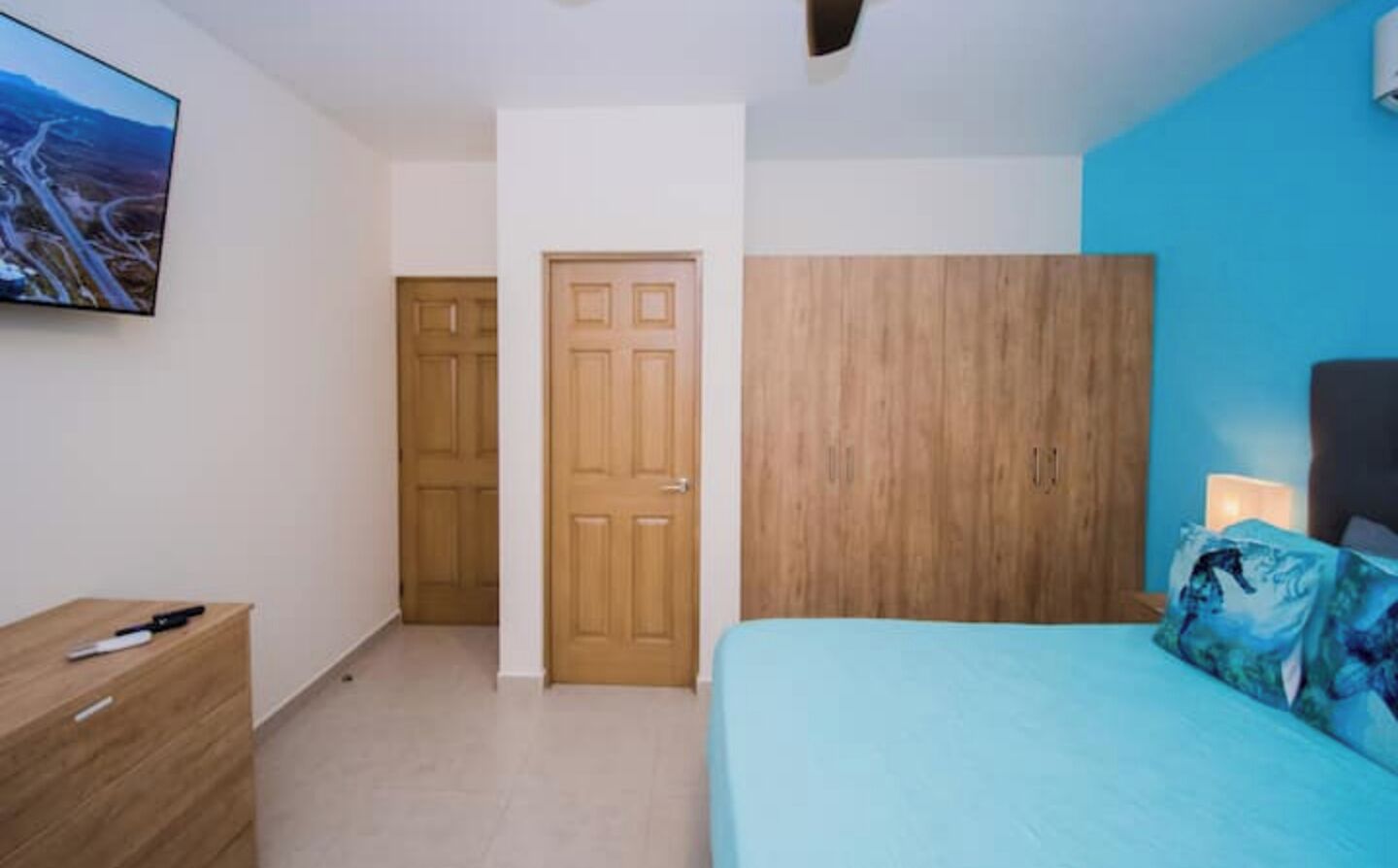 A bedroom with a bed, a wooden wardrobe, a desk with a chair, and a wall-mounted TV.