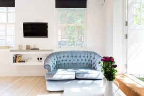 Un salon moderne avec un canapé bleu clair, un téléviseur à écran plat fixé au mur et une fenêtre donnant sur un paysage verdoyant.