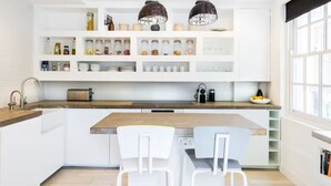 Une cuisine moderne avec des meubles blancs, un plan de travail en bois et des luminaires suspendus. On y trouve deux chaises blanches près de l’îlot central, ainsi qu’une variété de bocaux et d’ustensiles de cuisine sur les étagères.