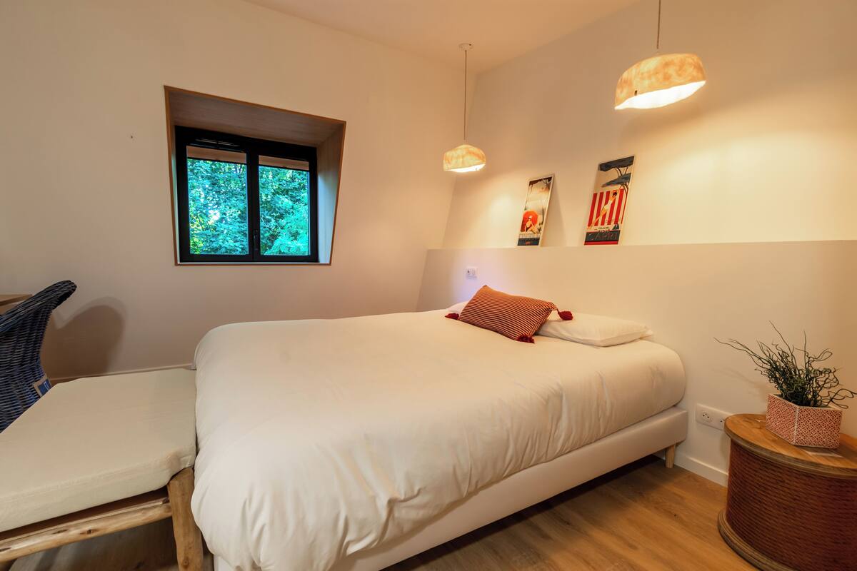 Une chambre avec un lit, une fenêtre, une lampe fixée au mur et une petite plante.